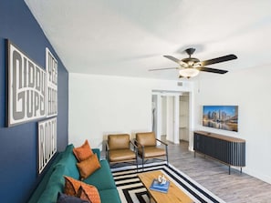 A living room with a sofa, two chairs, a coffee table, a ceiling fan, and a TV on a stand.