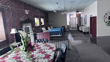 A dining area with a round table covered in a floral tablecloth, surrounded by chairs. There is a brick wall and a ceiling fan in the background.