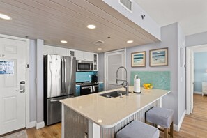 A modern kitchen with stainless steel appliances, a central island with seating, and a view of the adjacent living area. - Tower 3-405 Long Beach Resort Free Beach Chair Service! (Panama City Beach)