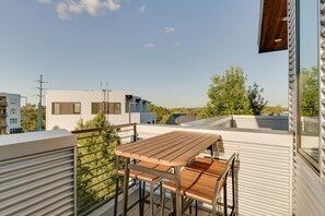 A rooftop terrace with a wooden table and chairs, metal railings, and a view of neighboring buildings and trees.