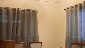 Ein Zimmer mit zwei Holzstühlen, einem kleinen Tisch und einer Couch. Vor den Fenstern hängen Vorhänge, und eine Pendelleuchte ist sichtbar.
