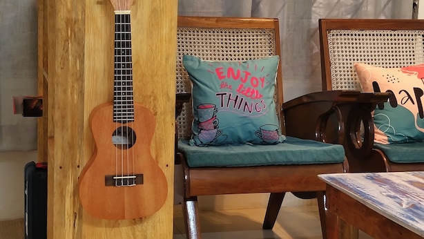 A wooden guitar stand with a guitar, a wooden chair with a cushion, and a table with a magazine. - zion backpackers hostel (Guwahati)