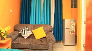 A compact living space with a sofa, two cushions, a small table with a vase of flowers, and a refrigerator. - Superhighway Haven, 2 Mins To SpurMall Kimbo-Ruiru,  EXIT 12&13 Thika Road (Kimbo, Ruiru)