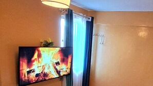 A hotel room with a bed, a TV mounted on the wall displaying a fire, a window with curtains, and a ceiling lamp. - Superhighway Haven, 2 Mins To SpurMall Kimbo-Ruiru,  EXIT 12&13 Thika Road (Kimbo, Ruiru)