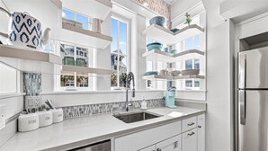 A modern kitchen with white cabinetry, a stainless steel refrigerator, and a window overlooking a residential area. - Cottage Charm, Easy Beach Access & Resort Amenities@Cinnamon Shore North (Port Aransas)