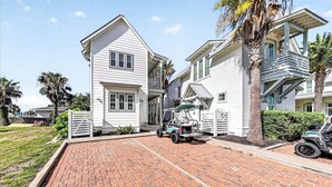 A two-story white building with a brick driveway, a covered outdoor seating area, and a vehicle parked in front. - Cottage Charm, Easy Beach Access & Resort Amenities@Cinnamon Shore North (Port Aransas)