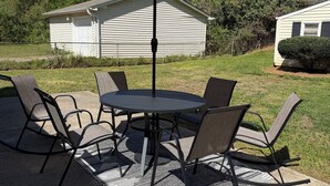 Um pátio externo com uma mesa redonda e seis cadeiras, um guarda-sol grande, e dois edifícios brancos ao fundo.