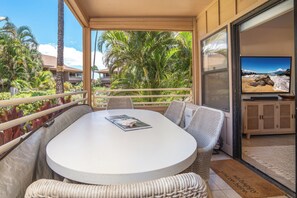 Un área de estar al aire libre con techo, con una mesa redonda y sillas de mimbre, con vista a una playa con palmeras y al océano.