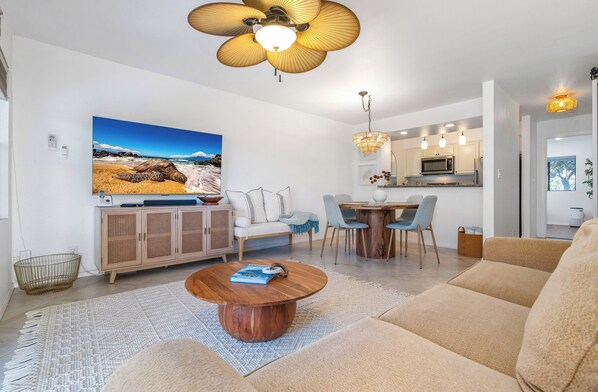 Una sala moderna con un televisor de pantalla plana grande, una mesa de centro redonda y un sofá. Hay una zona de cocina con una mesa de comedor y sillas.
