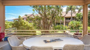 A covered outdoor seating area with wicker chairs and a round table, overlooking a landscaped garden and a building with multiple balconies.