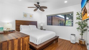 A bedroom with a wooden bed, a ceiling fan, a window with a view, a bedside table with a lamp, and a potted plant.