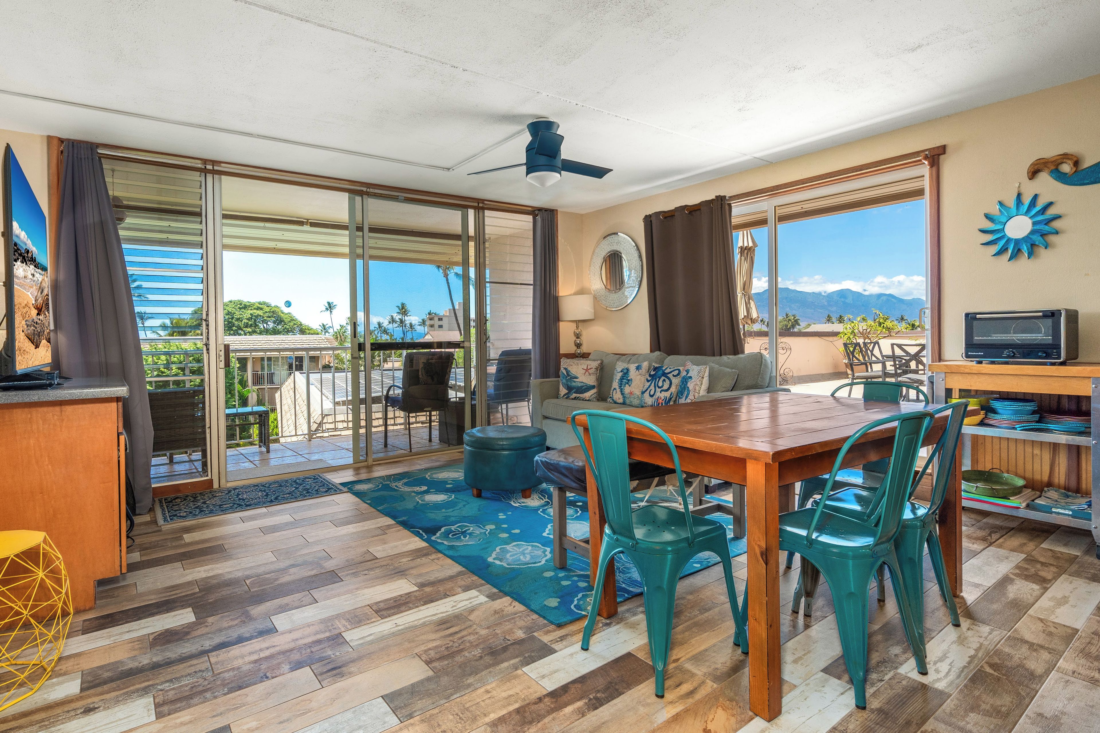 A spacious living area with a wooden dining table, teal chairs, a sofa, and a TV.