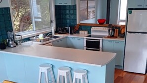 A kitchen with a central island, white stools, a refrigerator, and a window. - Ocean Blue - Linen,  WiFi, Kayo and Pet Friendly (Venus Bay)