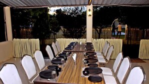 Una larga mesa de comedor dispuesta para un evento formal, rodeada de sillas blancas acolchadas, bajo un área cubierta con vistas a árboles y una valla.