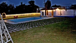 Una piscina con solárium empedrado, un pequeño espacio de estar al aire libre y un edificio con porche cubierto.