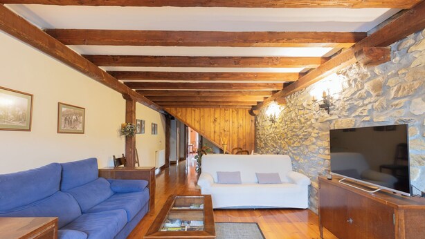 A cozy living room with a wooden ceiling, stone wall, and a large TV. - Zelaikoa Goikoa - baskeyrentals (Zestoa)