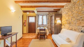 A cozy living room with a stone wall, wooden beams, a white sofa, a dining table with chairs, a television on a stand, and a fire extinguisher.