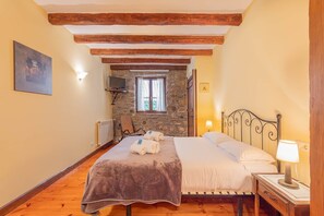 Un dormitorio con pared de piedra, vigas de madera, cama con manta, una silla, mesita de noche con lámpara y un cuadro en la pared.