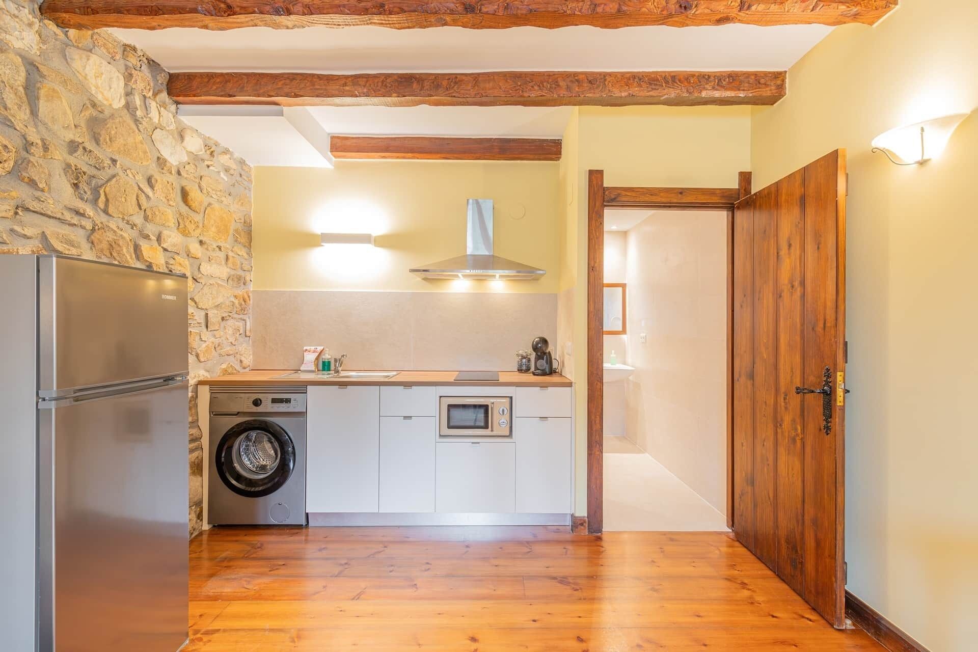 A compact kitchen with a stone wall, wooden beams, a washing machine, a built-in oven, and a wooden door.