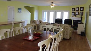 A dining area with a wooden table, white chairs, a glass pitcher, and a bowl of food. There is a sofa and a fireplace in the background. - Pelican Point #4 - Great Complex! Great Views! Cool off in the swimming pool! (Port Mansfield)
