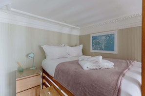 Un lit bien fait, avec des draps blancs, une couverture rose et une serviette pliée posée sur une tête de lit en bois.