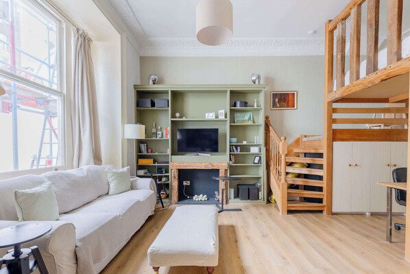 A living room with a sofa, ottoman, TV, wooden shelves, and a staircase.
