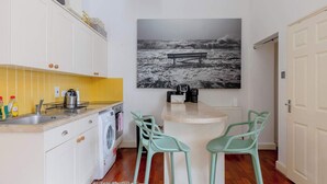 Une petite cuisine équipée d’un lave-linge, d’un évier et d’un plan de travail sur lequel se trouve une machine à café. On y trouve également deux chaises vertes et une grande photographie en noir et blanc accrochée au mur.