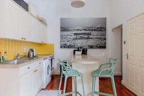 A compact kitchen with a washing machine, a sink, and a countertop with a coffee maker. There are two green chairs and a large black-and-white photograph on the wall.