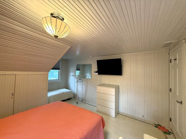 A bedroom with a sloped ceiling, a bed with an orange bedspread, a dresser, a TV mounted on the wall, and a window.