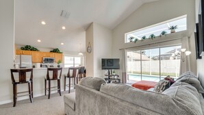 A spacious living area with a grey sofa, wooden chairs, a kitchen with wooden cabinets, a TV, and a large window overlooking a pool. - 15 Mi to Universal: Home w/ Pool (Kissimmee)