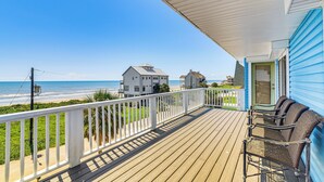 Eine Holzterrasse mit weißer Brüstung, die auf einen Strand blickt, ein blaues Gebäude und eine Bank.