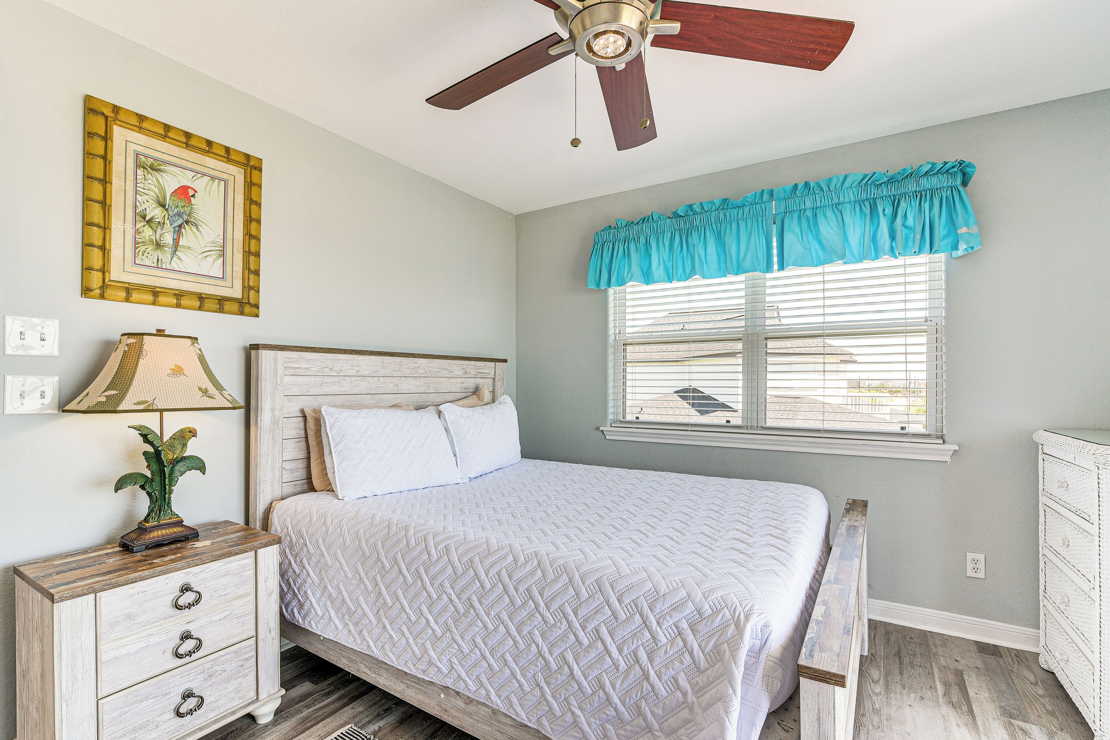 A bedroom with a bed, a nightstand, a lamp, a ceiling fan, a window with blinds, and a wall-mounted picture.