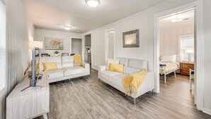 Un salon moderne avec un canapé blanc, une table basse et un téléviseur. Le sol est en bois et il y a un luminaire au plafond.