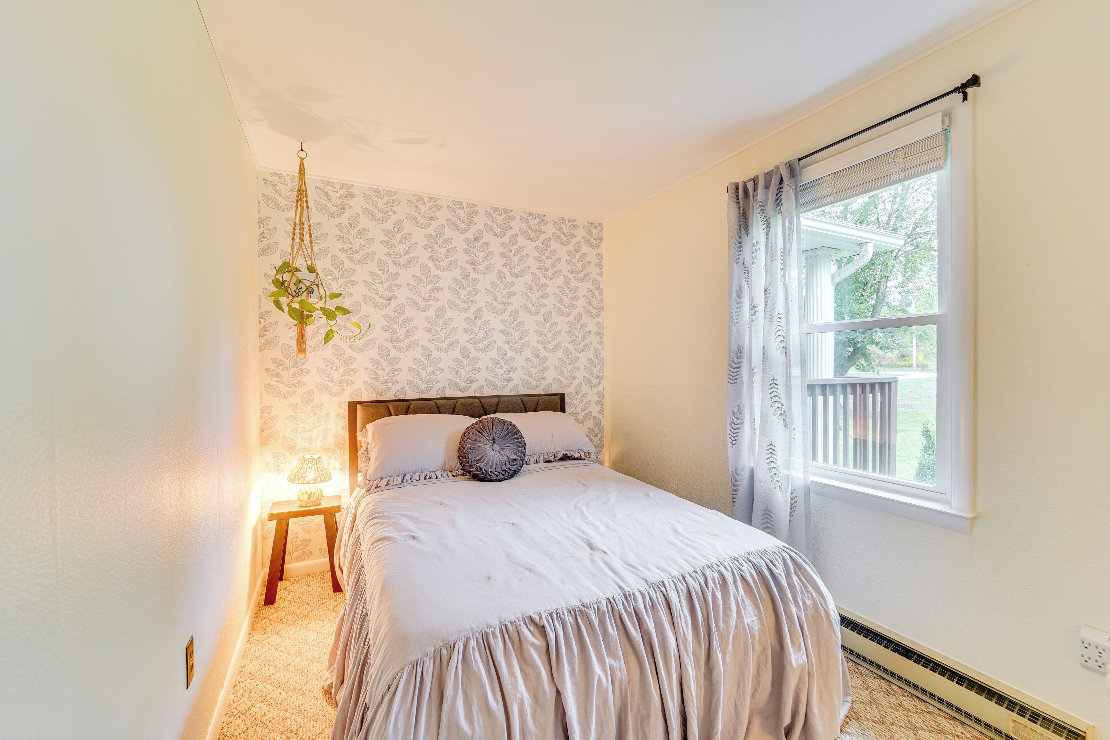 A bedroom with a bed, a bedside table with a lamp, a hanging macrame plant, a window with curtains, and a patterned wallpaper.