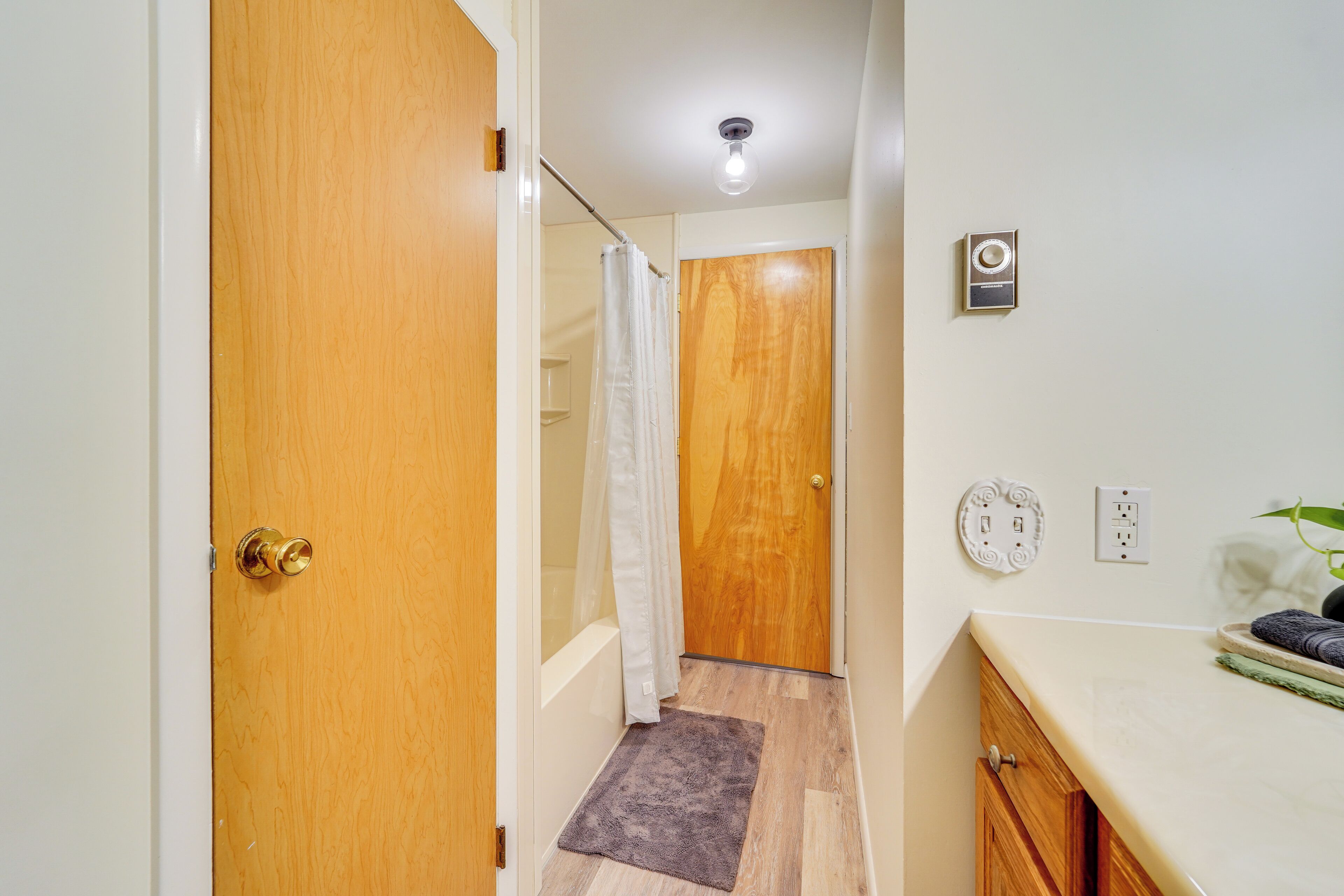 Un pasillo con una puerta de madera, una ducha con cortina blanca, una alfombra pequeña, una lámpara y un enchufe en la pared.