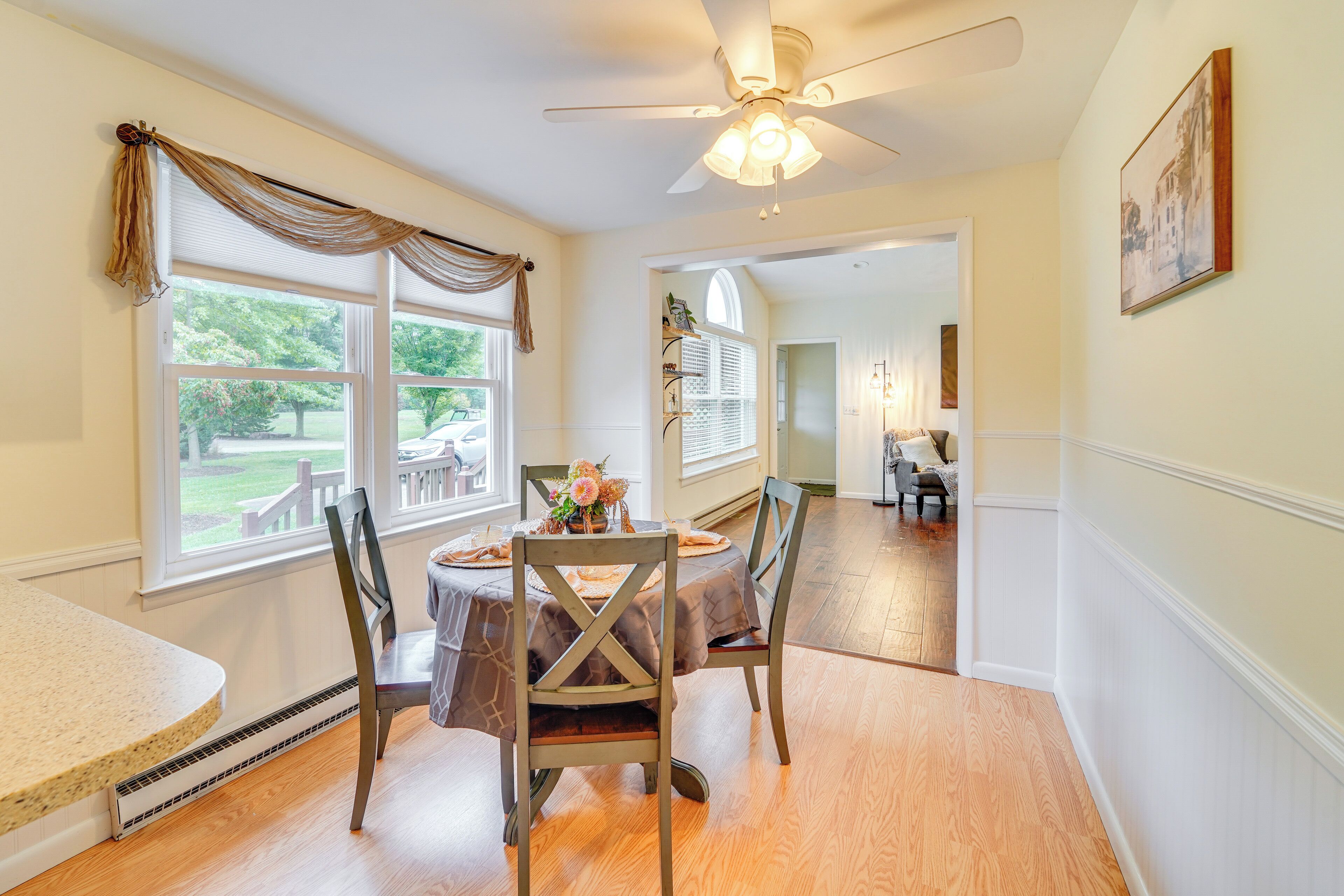 A dining area with a table set for four, wooden chairs, a ceiling fan, and large windows with curtains.