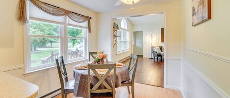 A dining area with a table set for four, wooden chairs, a ceiling fan, and large windows with curtains.