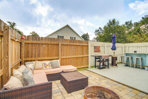 Ein Garten mit Holzzaun, einer Terrasse mit Rattan-Sofa und Hocker, einem Grill und einem kleinen Tisch mit einer Flagge.