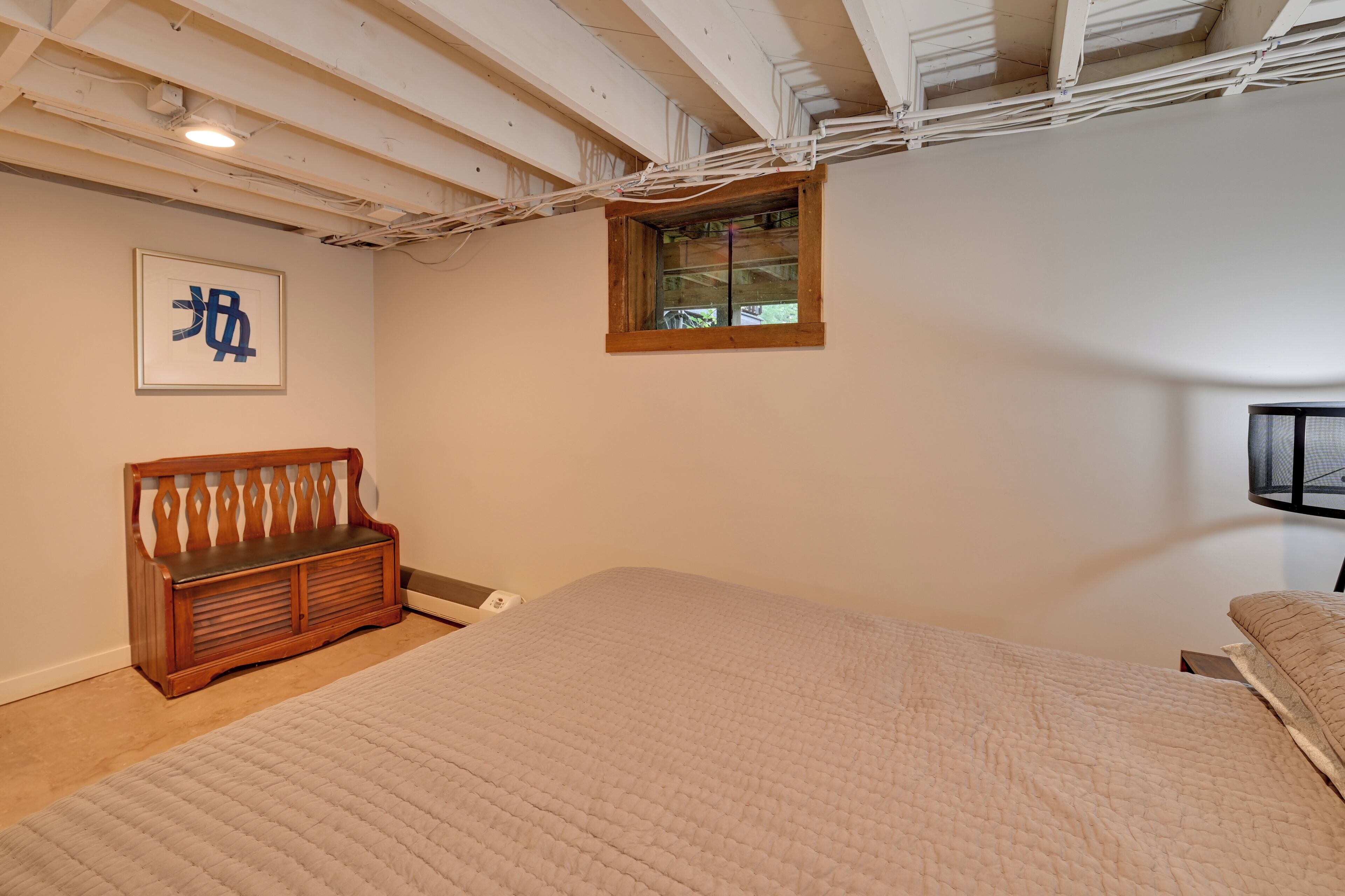 A bedroom with a large bed, a wooden bench, a small window, and a framed picture on the wall.