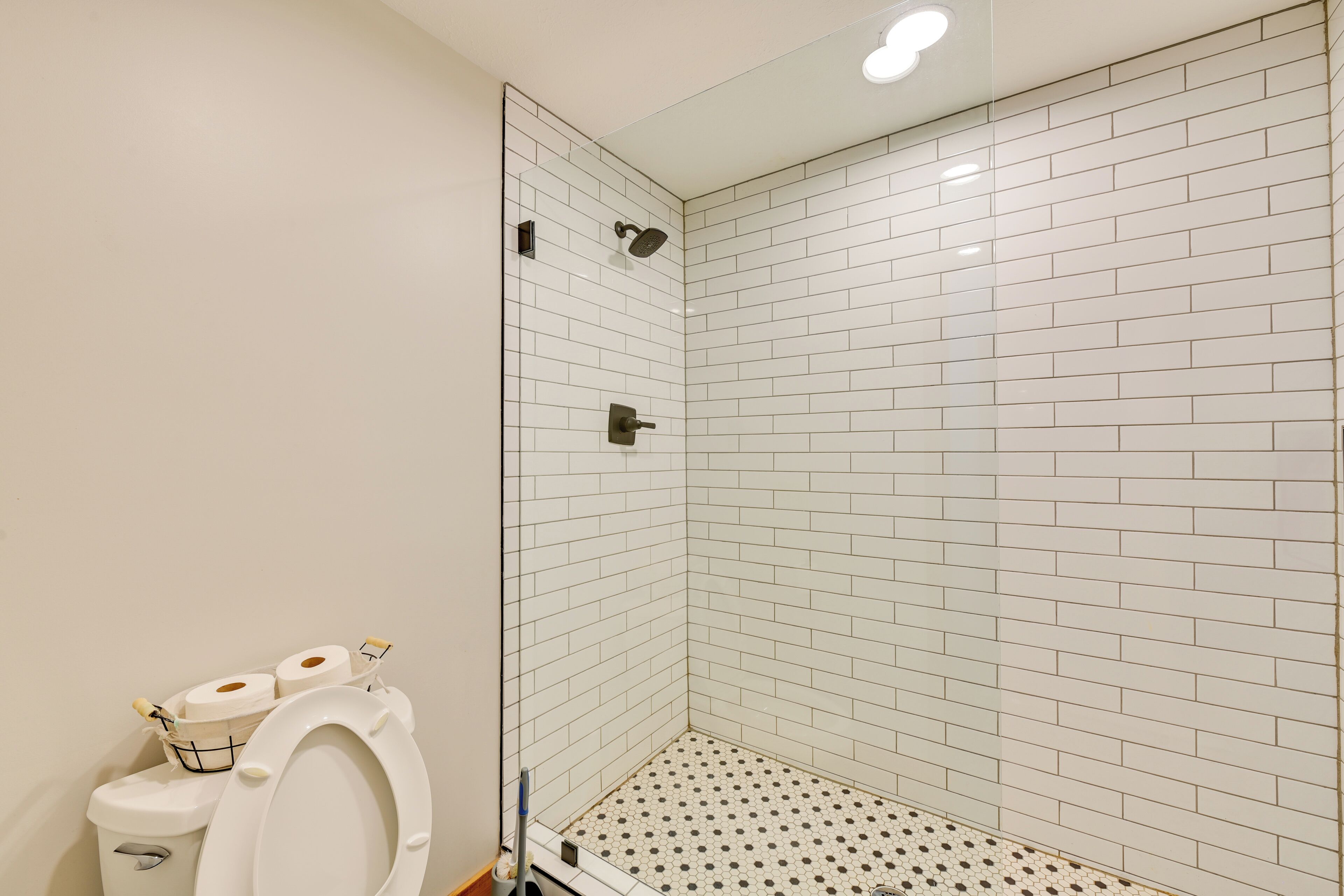 A bathroom with white tiled walls, a shower area, and a toilet with toilet paper and a toothbrush.