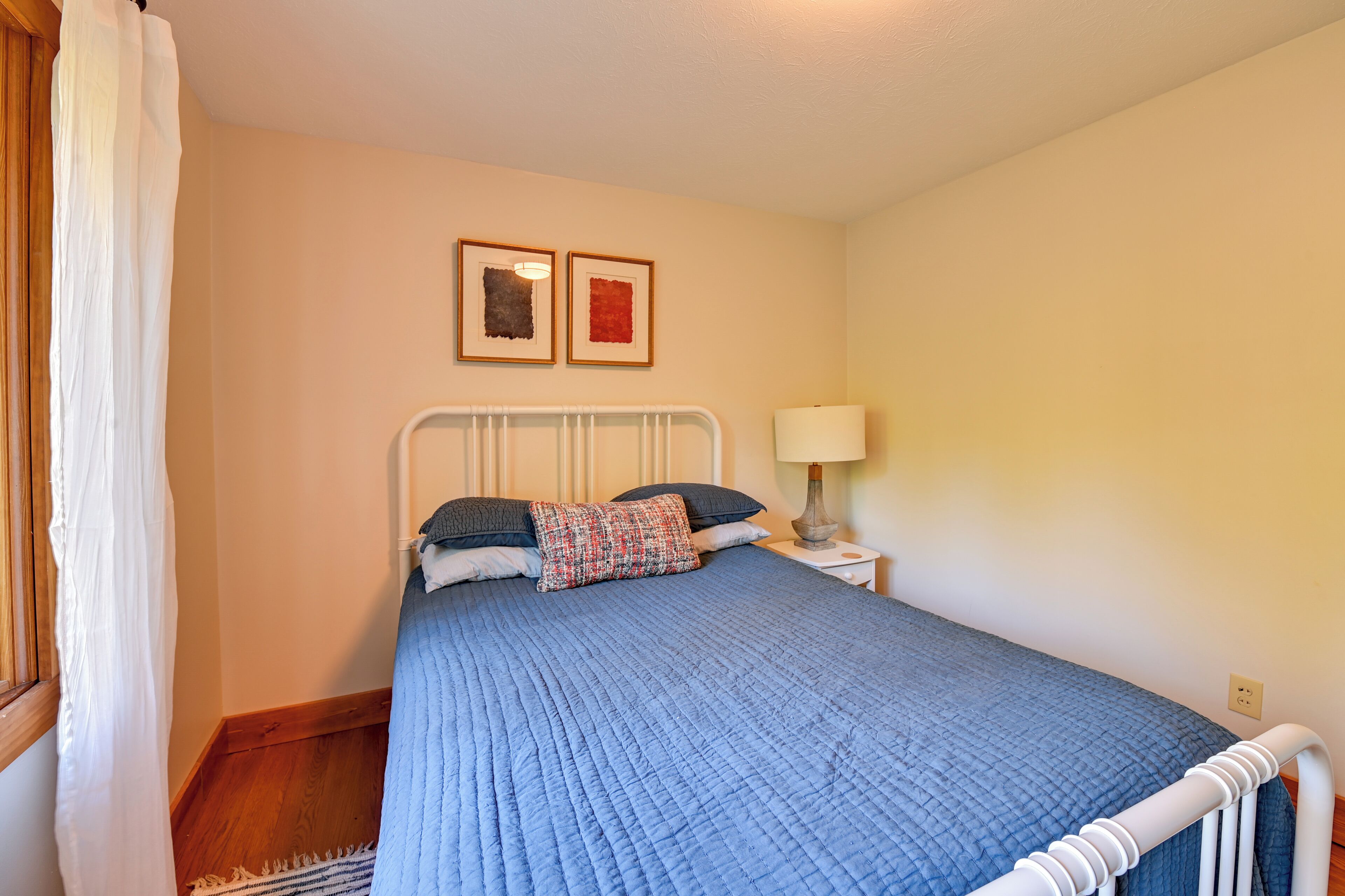 A bedroom with a bed, white headboard, bedside table with a lamp, and two framed pictures on the wall.