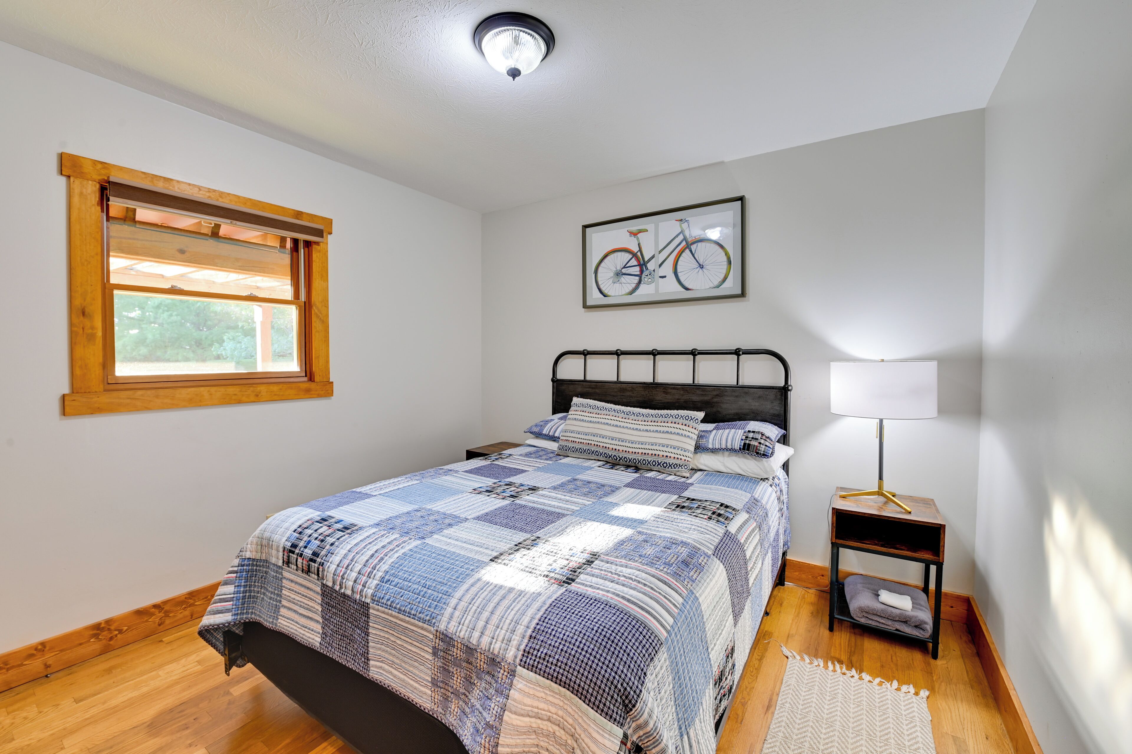 A bedroom with a bed, a window, a wall-mounted bicycle artwork, a bedside lamp, and a small wooden nightstand.