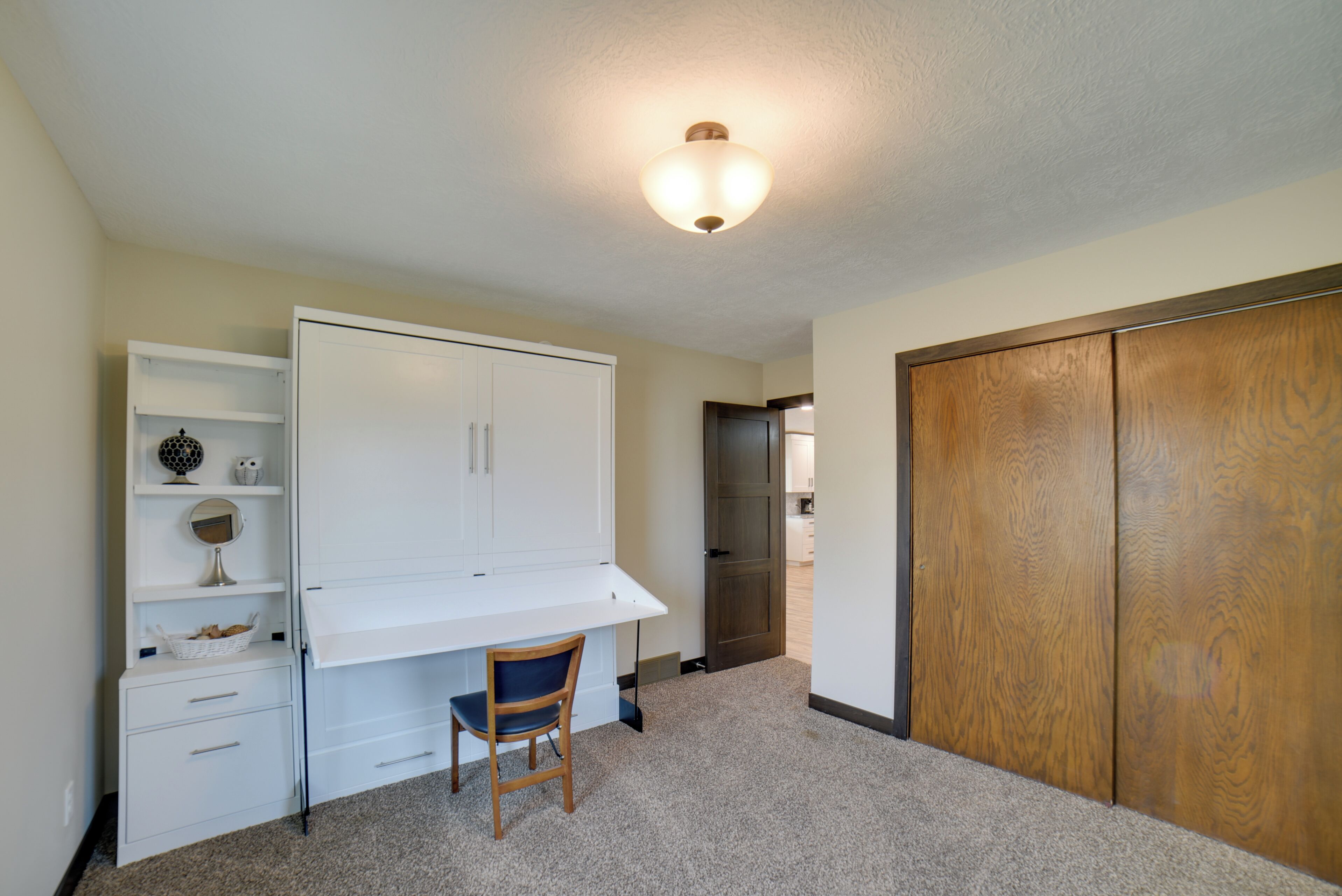 Une pièce avec un bureau blanc, une chaise, une armoire en bois et un luminaire fixé au plafond.