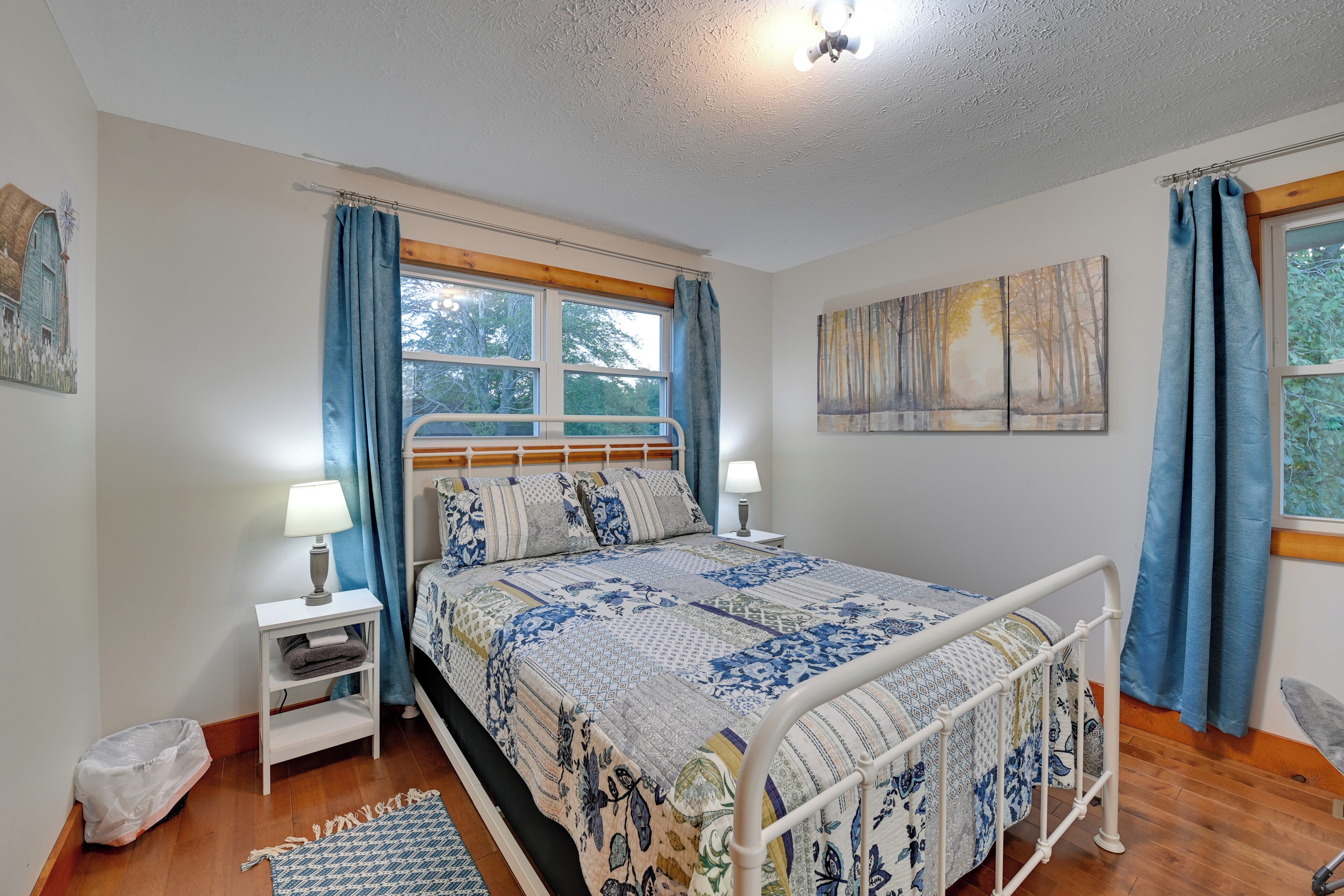 A bedroom with a bed, two bedside tables, a window with curtains, a painting on the wall, and a wooden floor.