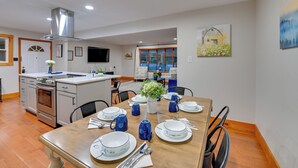 Een moderne keuken met roestvrijstalen apparatuur, een eethoek met een houten tafel gedekt voor vier personen, en een groot raam met uitzicht op een huis.