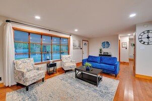 Una sala de estar con un ventanal grande, un sofá azul, dos sillones estampados, una mesa de centro y un reloj en la pared.
