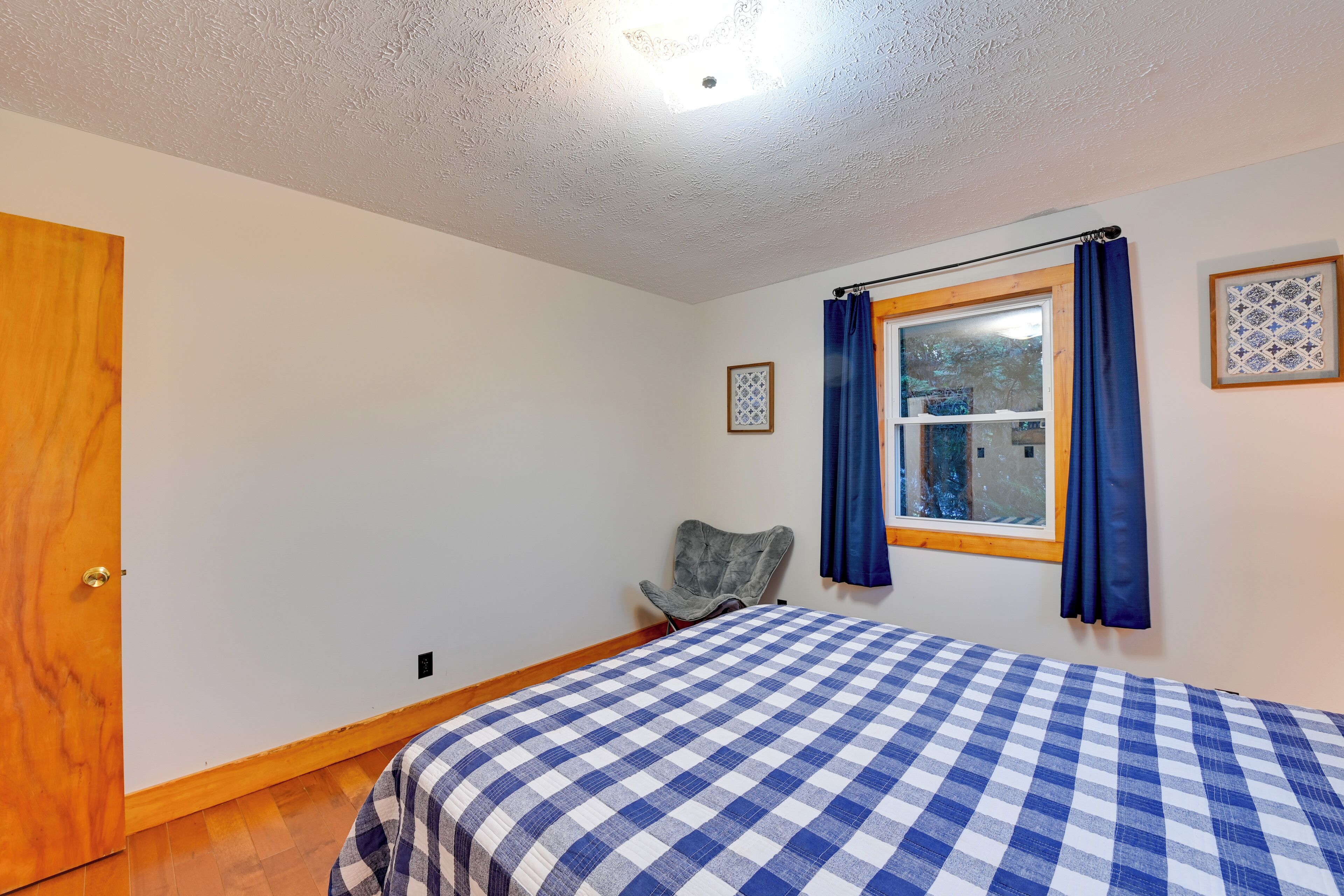 Une chambre avec une couvre-lit à carreaux, une porte en bois, une fenêtre avec des rideaux bleus et un tableau encadré au mur.