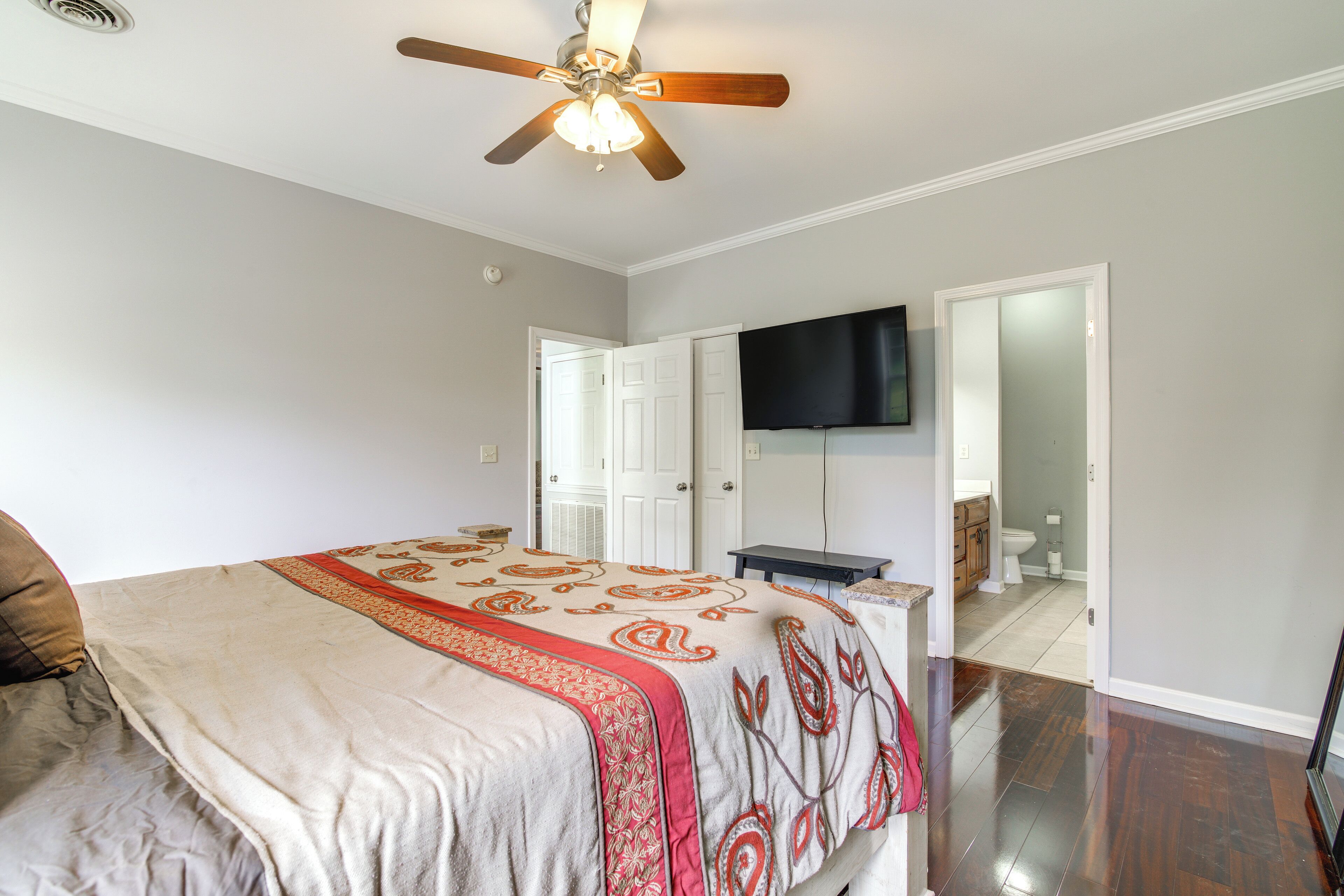 A bedroom with a bed, a ceiling fan, a television mounted on the wall, and a bathroom visible through an open door.