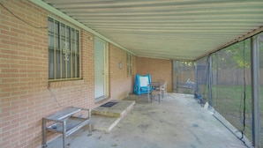 Ein überdachter Patio-Bereich mit Backsteinmauer, einer Tür, einem Fenster mit Gitter und einem blauen Stuhl sowie einem Tisch.