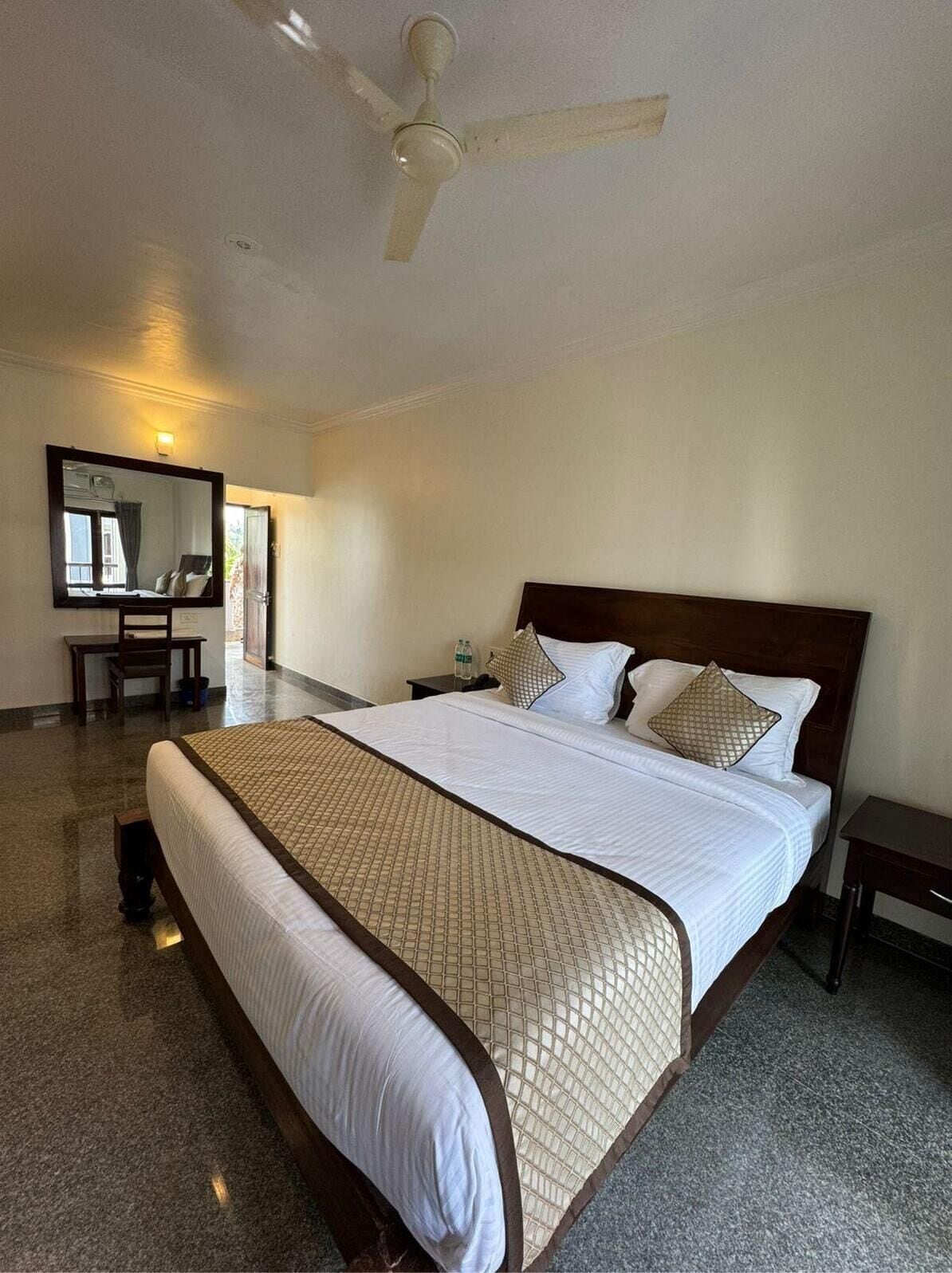 A hotel room with a large bed, a ceiling fan, a bedside table, a nightstand, a mirror, and a view of the outside.